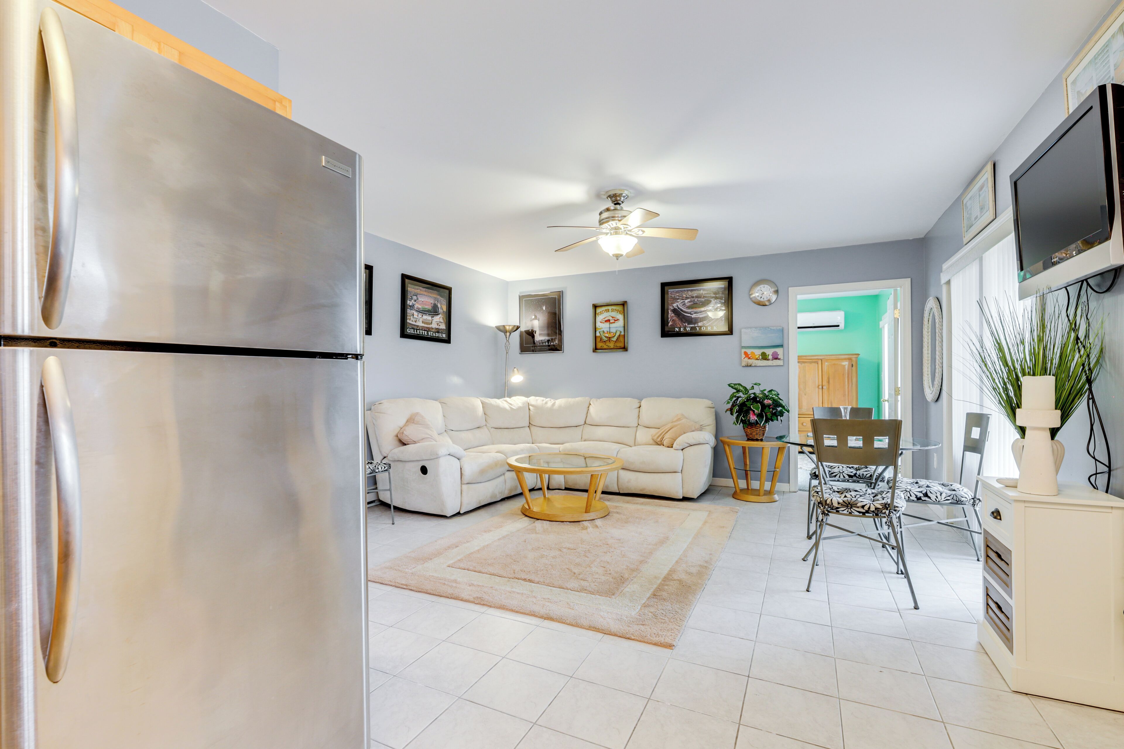 A modern living room with a sofa, coffee table, and chairs. There is a refrigerator, a ceiling fan, and a TV mounted on the wall.