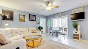 A modern living room with a beige sofa, a glass coffee table, a ceiling fan, and a flat-screen TV mounted on the wall. - Walk to Eastern Point Beach: Apt w/ Water View! (Groton)