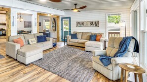 A spacious living room with two sofas, a coffee table, and a rug. There is a ceiling fan, a window with a view of trees, and a sign that reads "FUN IN THE SUN."