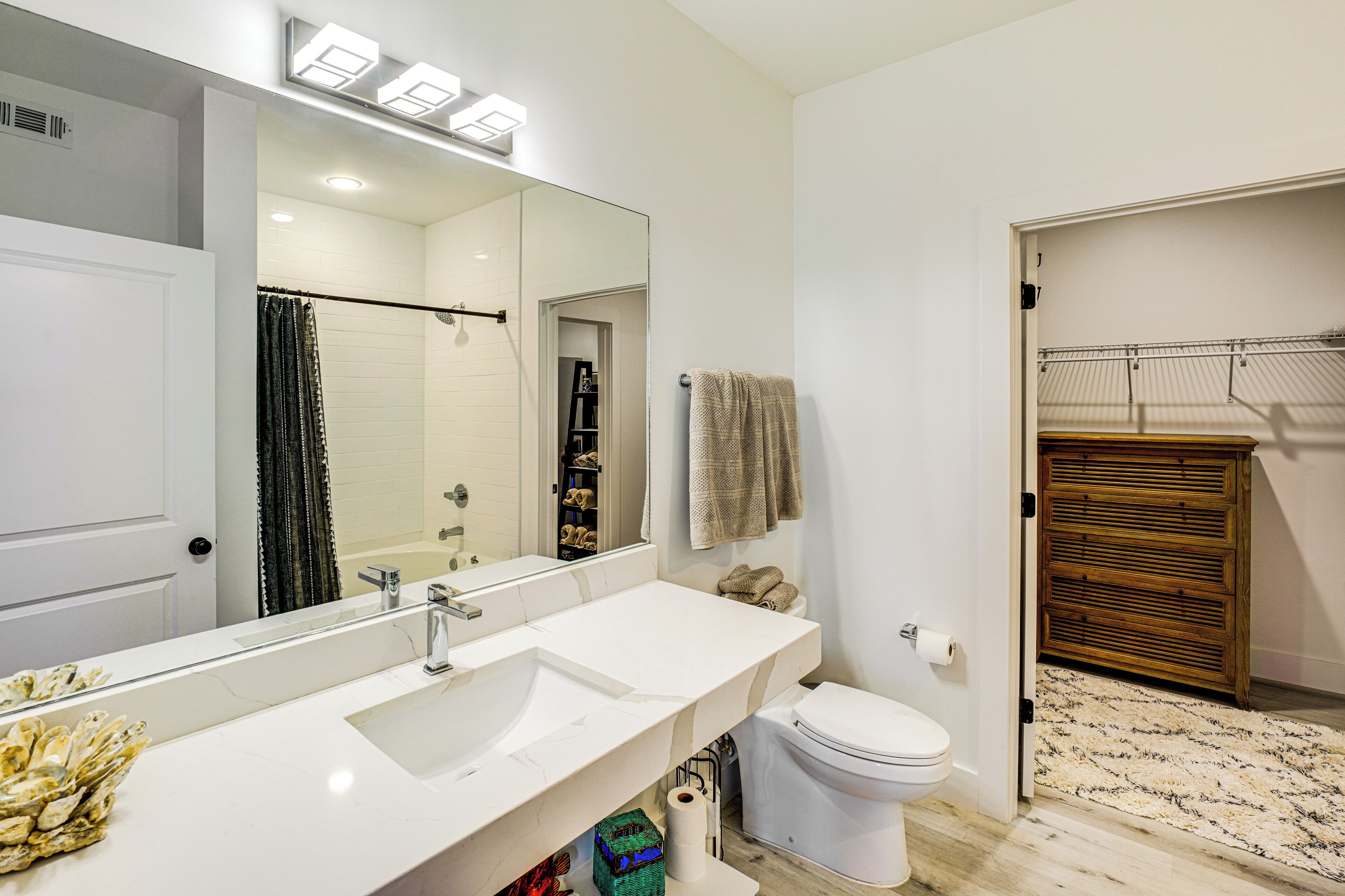 A modern bathroom with a large mirror, white sink, and toilet. There is a shower area with a glass door and a wooden storage unit.