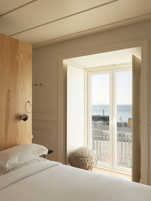 Un dormitorio con una cama, un armario de madera, un otoman redondo y un balcón con vista al mar.