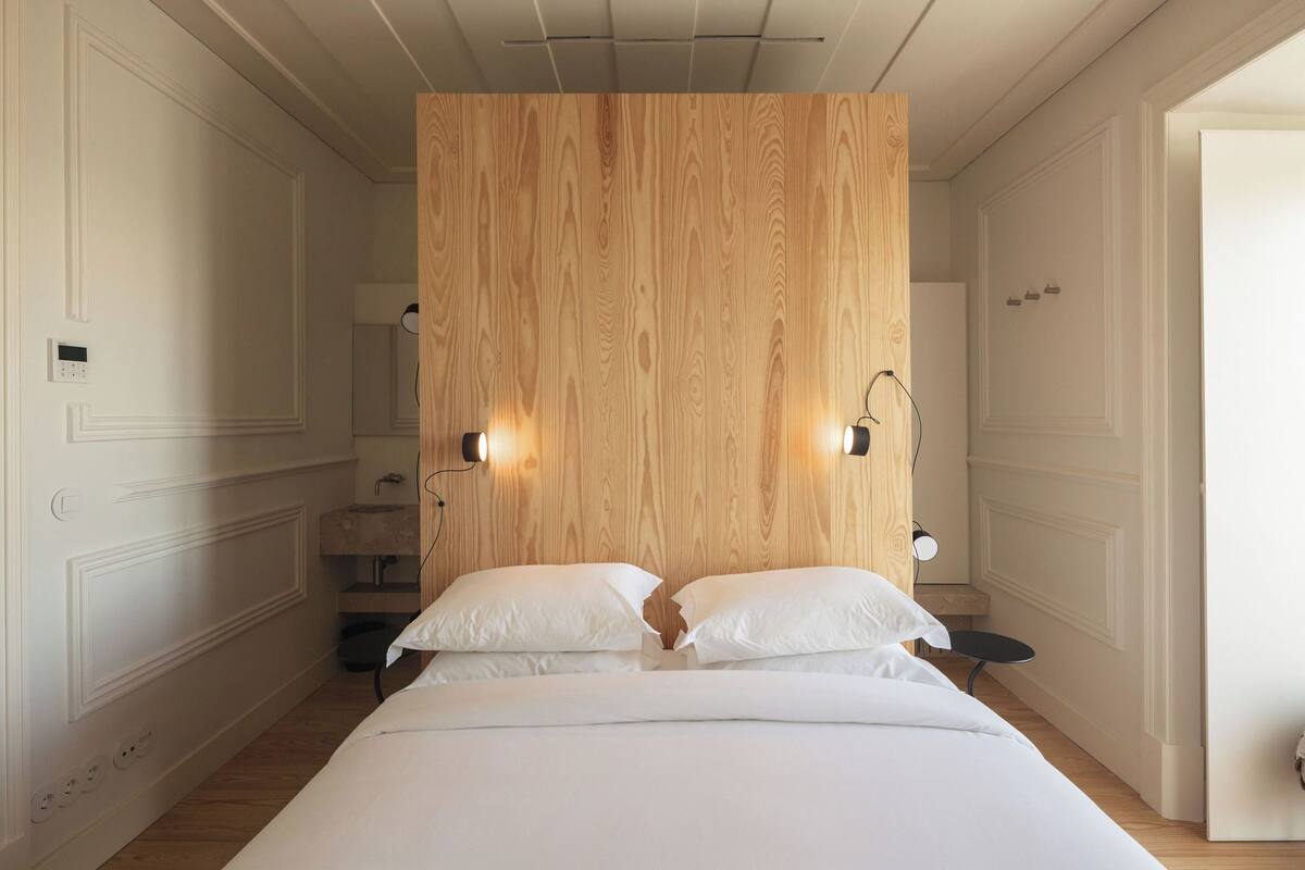 A bedroom with a wooden headboard, two bedside lamps, a bed with white bedding, and a small round table.