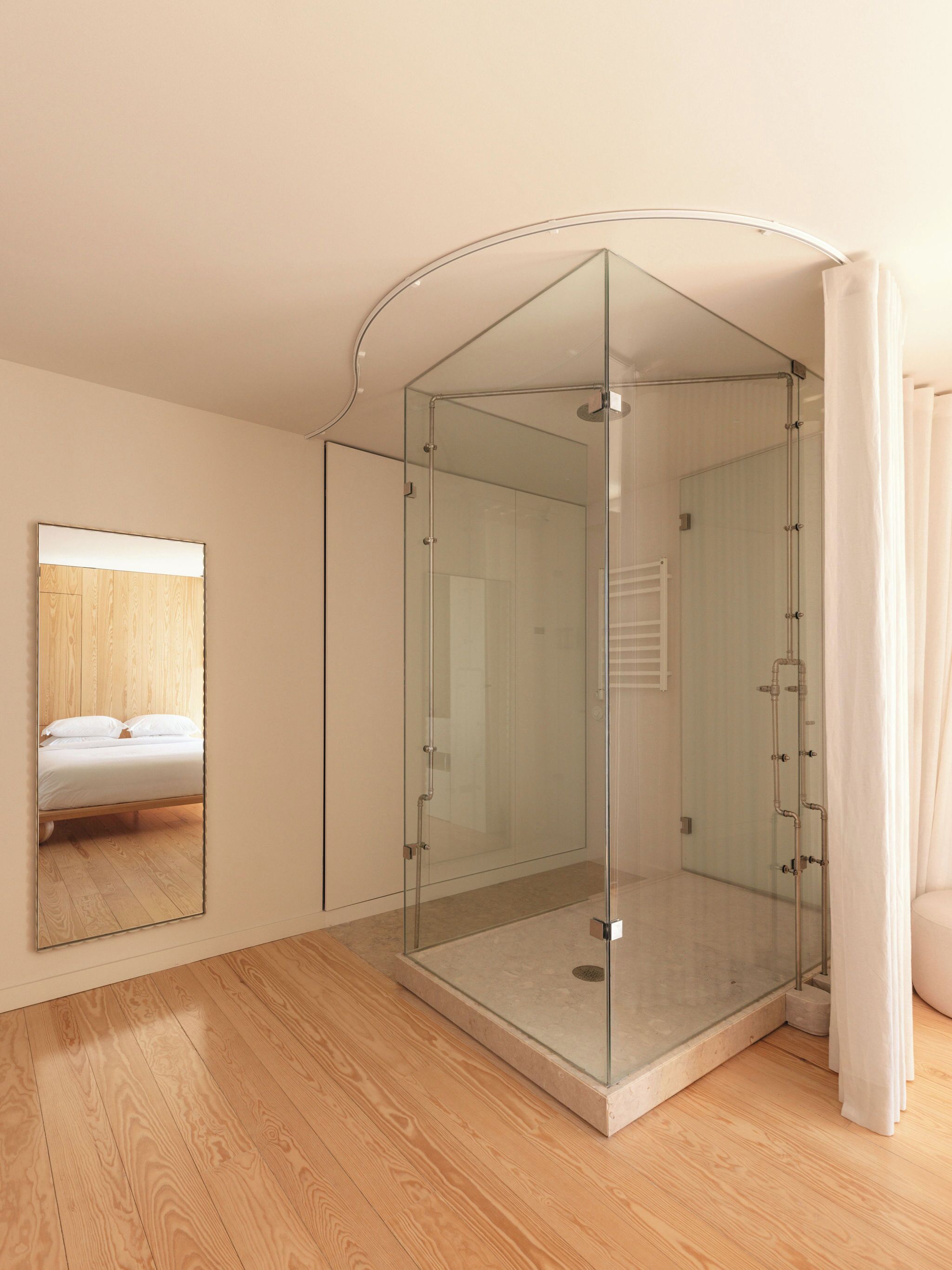 Un bagno moderno con cabina doccia in vetro, pavimento in legno e uno specchio che riflette un letto.