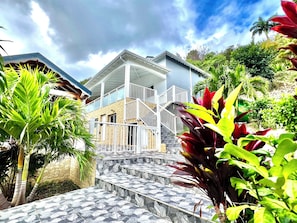 A two-story building with a balcony, surrounded by tropical plants.