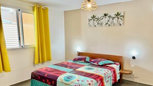 Una habitación con una cama, cortinas amarillas, una lámpara de pared y un cuadro con motivos tropicales.