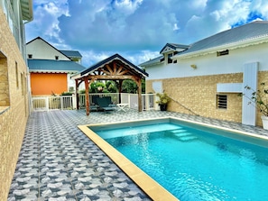 A swimming pool with a tiled deck, a covered seating area, and a stone wall.