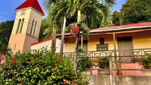 A building with a clock tower, a sign reading "Honeymoon Police", and a palm tree with red flowers. - Zandoli Apartment. Bas Vent Estate. (Deshaies)