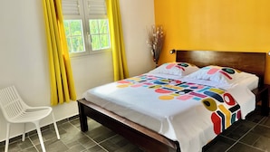 A room with a bed, a chair, a window with curtains, and a wall painted yellow. - Zandoli Apartment. Bas Vent Estate. (Deshaies)