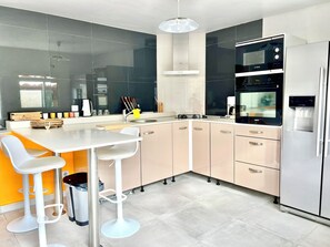 A modern kitchen with a built-in oven, refrigerator, and a dining area with bar stools. - Colibri Apartment. Bas Vent Estate. (Deshaies)