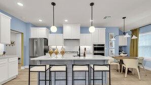 A modern kitchen with white cabinets, a central island with bar stools, and pendant lighting. - The Bella Muse - 3-Bedroom House W/ Hot Tub close to Bike Trails (Bella Vista)