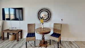 A living room with a round wooden table, two wicker chairs with blue cushions, a wall-mounted TV, and a decorative mirror.