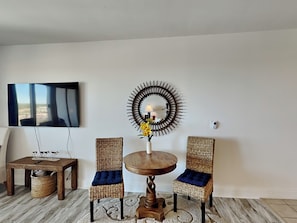 Una sala de estar con una mesa redonda de madera, dos sillas de mimbre con cojines azules, un televisor montado en la pared y un espejo decorativo.