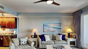 A living room with two sofas, a coffee table, and a ceiling fan. There is a kitchen area with wooden cabinets and a heart-shaped decoration on the wall.