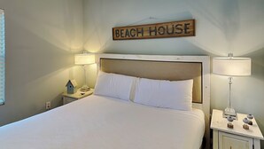 A bedroom with a bed, two bedside tables, lamps, a wall-mounted sign reading "BEACH HOUSE", and a small birdhouse on one of the nightstands.