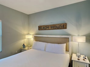 Un dormitorio con cama, dos mesitas de noche, lámparas, un cartel en la pared que dice 'BEACH HOUSE' y un pequeño comedero para pájaros sobre una de las mesitas.