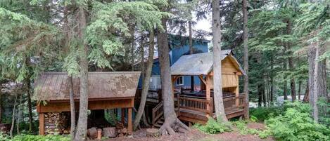 A cabin with a wooden deck and a porch, surrounded by tall trees in a forest.