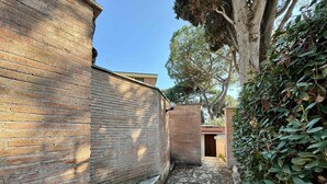 A narrow brick pathway leading to a building with a wooden door, surrounded by greenery and trees.