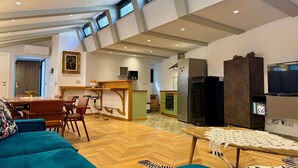A spacious living area with a wooden herringbone floor, a teal sofa, a dining table with chairs, a kitchen area with a refrigerator and oven, and a television on a stand.