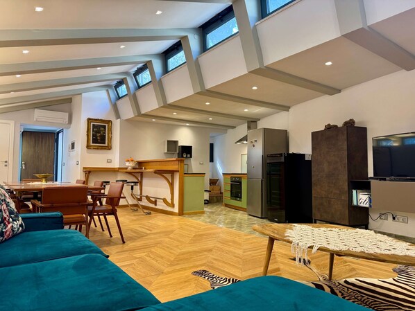 A spacious living area with a wooden herringbone floor, a teal sofa, a dining table with chairs, a kitchen area with a refrigerator and oven, and a television on a stand.