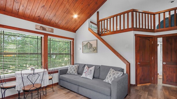 A living room with a grey sofa, a dining table with chairs, a staircase, and a large window with blinds. - Charming Tiny House Near Lake & Springs (Lampasas)