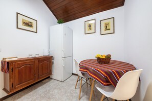 A compact kitchen with a white refrigerator, wooden cabinets, a striped tablecloth-covered table, and two framed pictures on the wall. - Pereira Place Cottage (São Vicente)