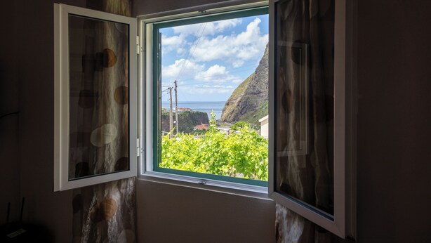 A room with a window offering a view of a green landscape and a distant coastline. - Pereira Place Cottage (São Vicente)