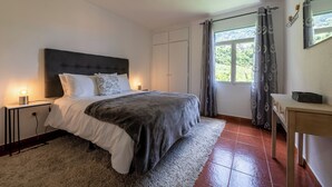 A bedroom with a bed, bedside tables, a nightstand, a window with curtains, and a view of greenery outside. - Pereira Place Cottage (São Vicente)