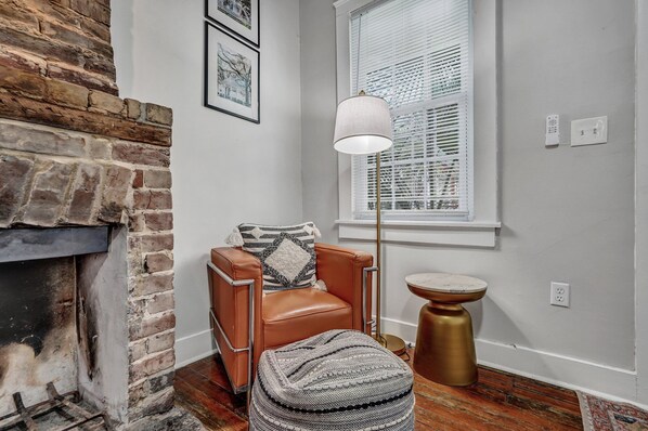 A room with a brick fireplace, a leather armchair with a throw pillow, a floor lamp, and a round table.