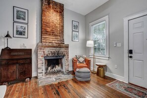 A room with a brick fireplace, wooden flooring, a white door, and a window with blinds.