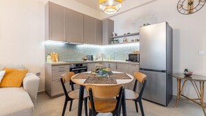 A modern kitchen with a dining area, featuring a round table, chairs, a refrigerator, and a clock on the wall. - Petra - Stylish Apartment (Hersonissos)