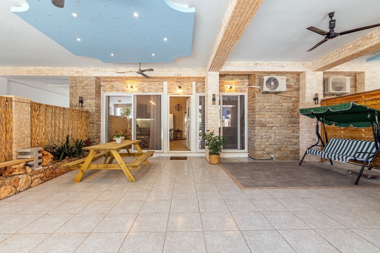 A spacious patio area with a wooden table and benches, a ceiling fan, and a green canopy.