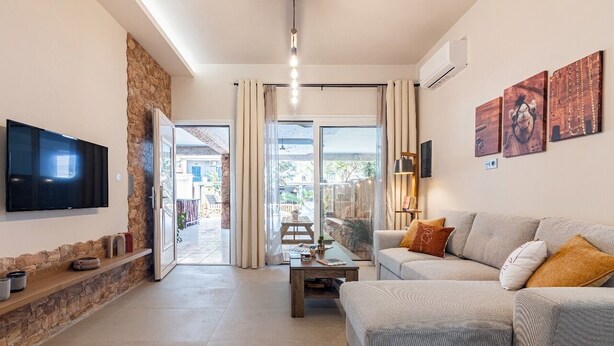 A modern living room with a flat-screen TV, a stone accent wall, a grey sofa with cushions, and a large window offering a view of the outdoors. - Petra - Stylish Apartment (Hersonissos)