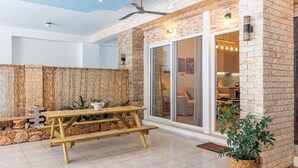 A modern outdoor patio with a wooden picnic table, stone walls, and a glass door leading to an interior space. - Petra - Stylish Apartment (Hersonissos)