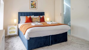 A neatly made bed with a blue headboard, white linens, and orange accents. There is a small bedside table with a lamp and a decorative item. A colorful abstract painting hangs above the bed.