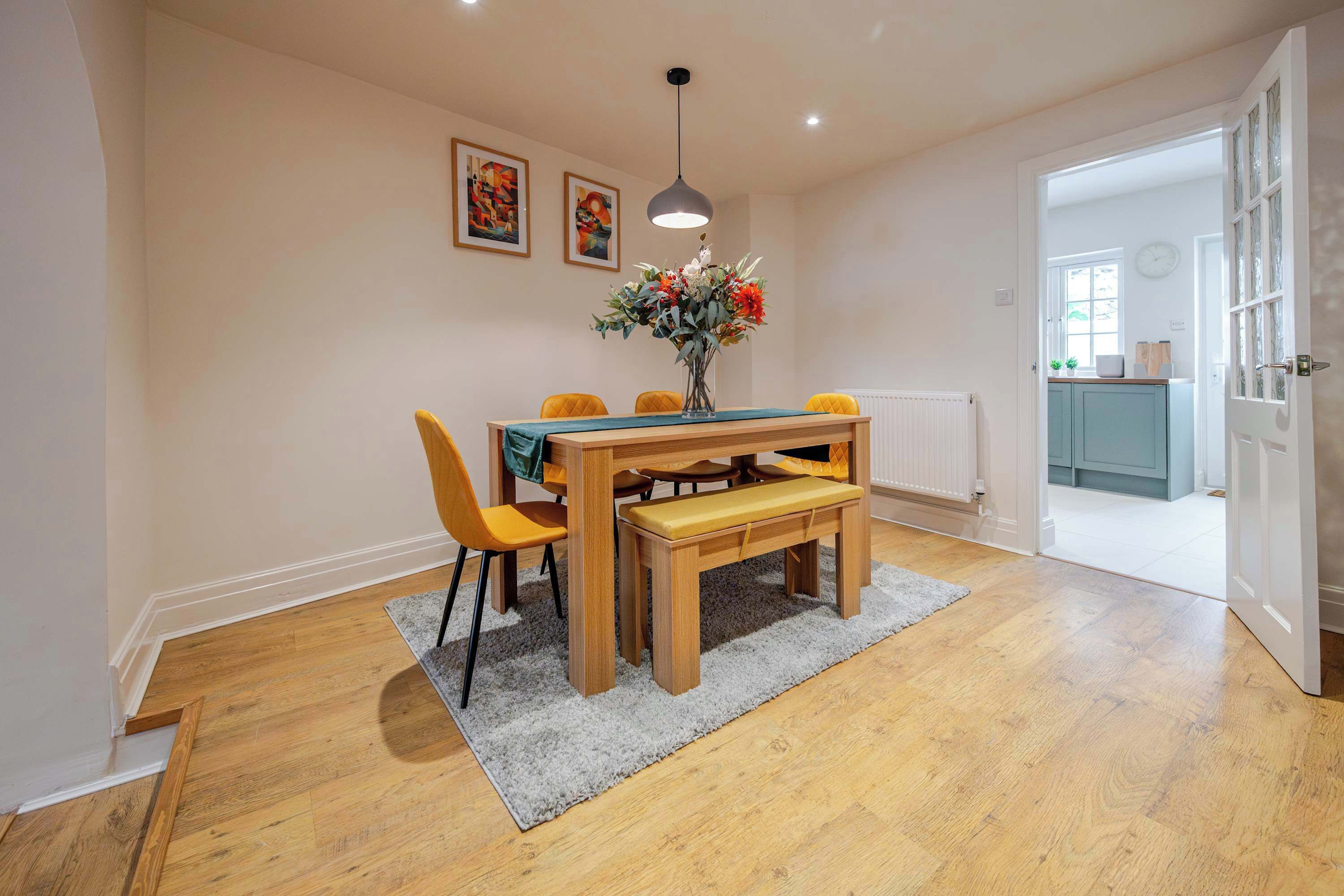 Ein Essbereich mit einem Holztisch, vier gelben Stühlen, einer Bank und einem Blumenstrauß. Unter dem Tisch liegt ein grauer Teppich, und der Boden ist mit Holz ausgelegt.