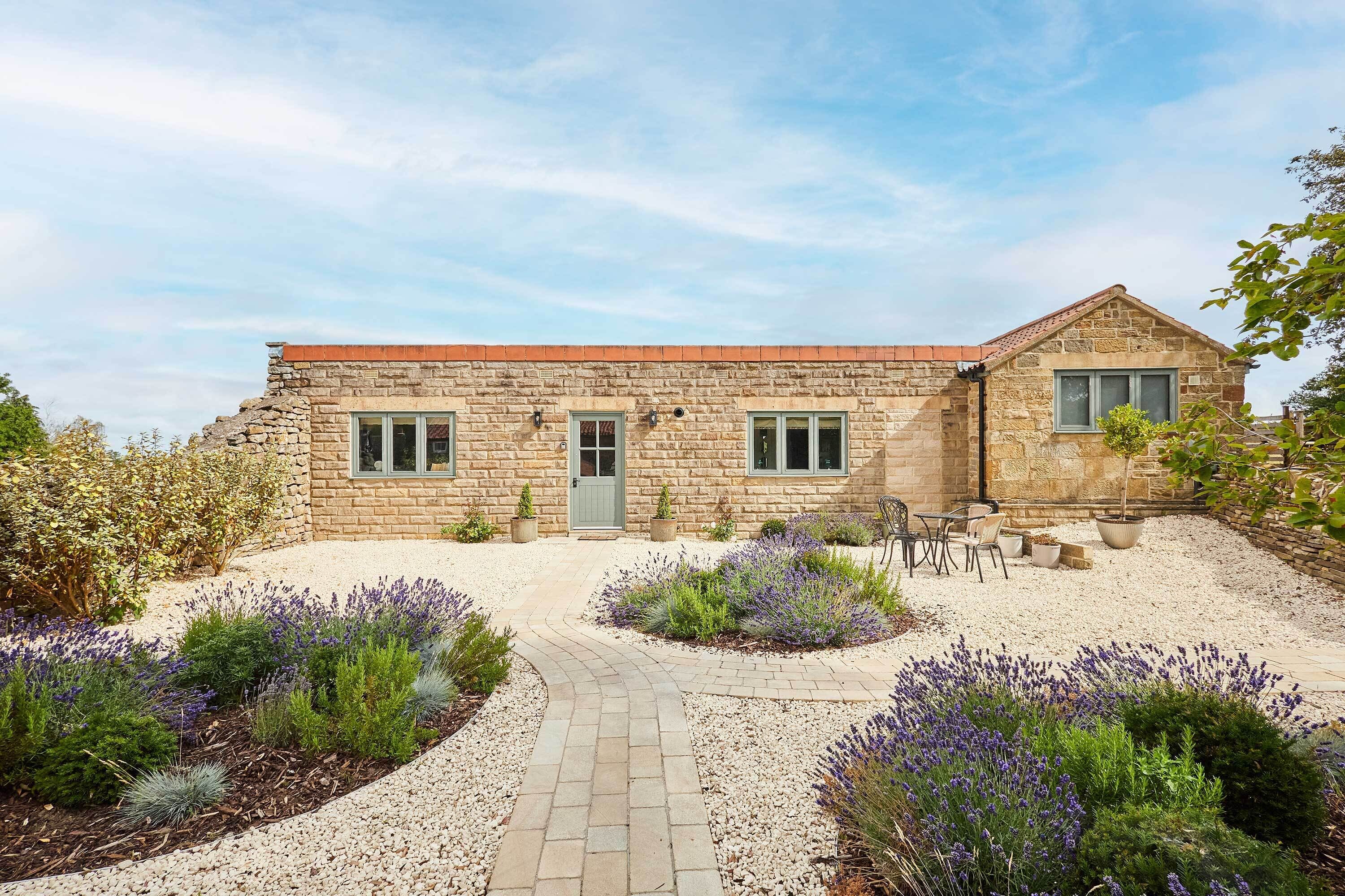 Un bâtiment en pierre avec une entrée pavée, un petit jardin agrémenté de lavande et des sièges à l’extérieur.
