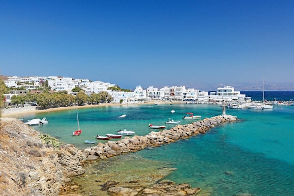 Un pueblo costero con edificios blancos, aguas turquesas cristalinas y un rompeolas rocoso.