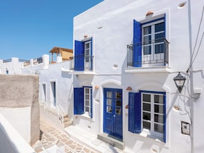 A white building with blue shutters and a balcony, a stone staircase, and a small window with a light fixture. - Marouso Villa - Paros Seaview Getaway (Marpissa)