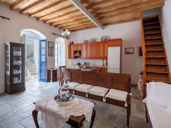 A spacious room with a wooden ceiling, a dining area with a table set for two, a kitchen with wooden cabinets, and a staircase leading to an upper level.