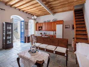 Una habitación amplia con techo de madera, un comedor con mesa dispuesta para dos personas, una cocina con armarios de madera y una escalera que sube a un nivel superior.