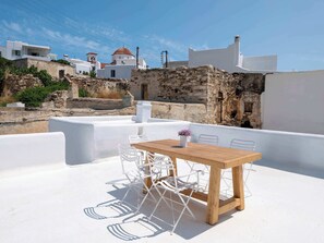 Una terraza en la azotea con una mesa de madera y sillas blancas, con vistas a un edificio encalado con tejados de tejas rojas y muros de piedra.