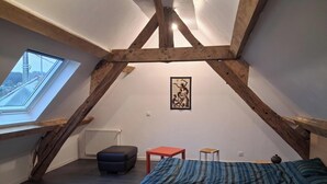 Ein Schlafzimmer mit schrägen Wänden, Holzbalken, einem Oberlicht, einer Heizung, einem Bett mit gestreifter Decke, einem kleinen Tisch und einem Stuhl.