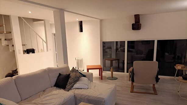 Ein modernes Wohnzimmer mit einem weißen Sofa, einem Holztisch und einem Sessel. Links befindet sich eine Treppe und rechts ein großes Fenster.