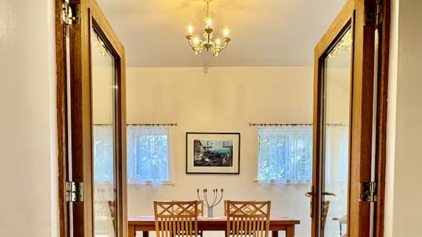 A dining area with a wooden table and chairs, a chandelier, and two windows with curtains.