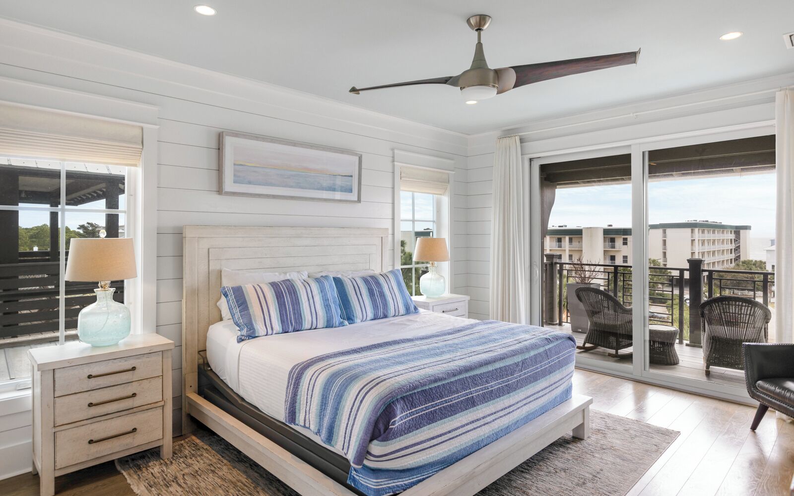 A bedroom with a large bed, a ceiling fan, and a balcony with wicker chairs.