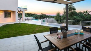A modern outdoor dining area with a view of the sunset, a pool, and a well-maintained lawn. - Villa Estel | Estel Residences (Chania)