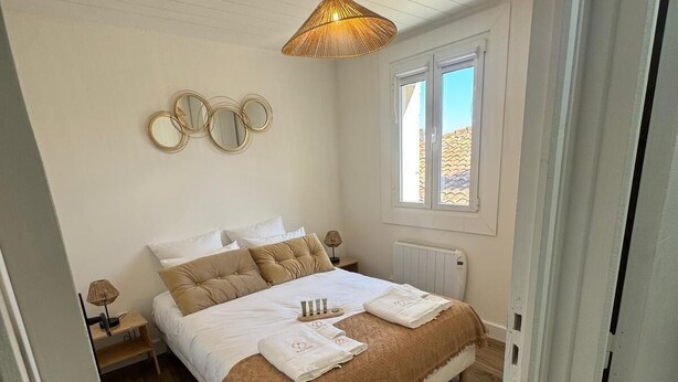 A bedroom with a wooden floor, a bed with a brown and white bedspread, a bedside table with a lamp, and a window with a view of buildings. - Résidence de la Place (Pont-du-Château)