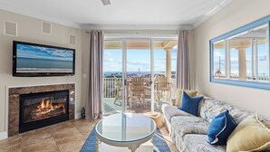 A living room with a fireplace, a television, a balcony with outdoor furniture, and a large window offering a view of the ocean. - Belmont Towers 710 by Seaside Vacations & Sales (Ocean City)
