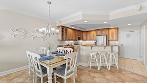 A spacious kitchen with wooden cabinets, a central island with stools, and a dining area with a glass table and white chairs. - Belmont Towers 710 by Seaside Vacations & Sales (Ocean City)