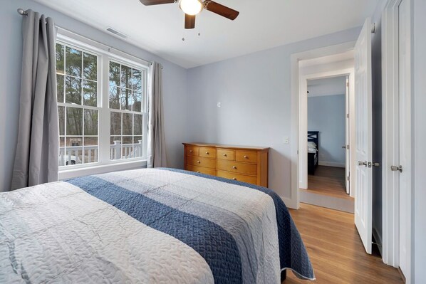 A bedroom with a large bed, a wooden dresser, a ceiling fan, and a view of a hallway.
